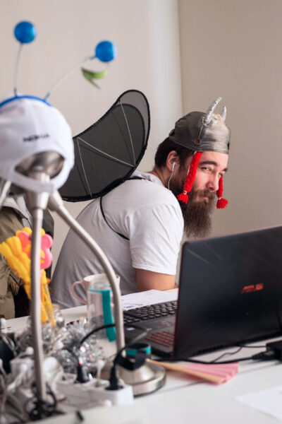 Man dressed in a whimsical costume with wings and a fun hat, sitting at a desk in front of a laptop, looking amused.