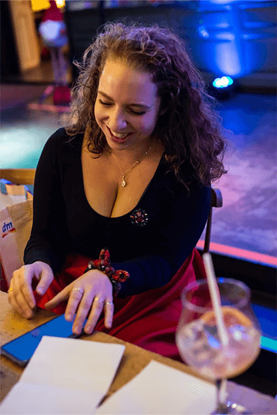 Woman sitting at a table, smiling while handling papers, with a drink nearby in a dimly lit setting.