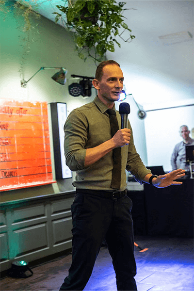 Man speaking into a microphone in a brightly lit room, wearing a green shirt and tie.