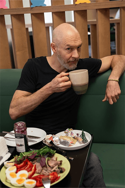 Man in a black shirt enjoying a large mug of coffee at a table filled with a variety of breakfast foods.