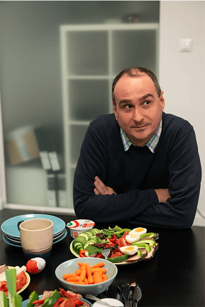 Man in a dark sweater sitting at a table with a variety of fresh vegetables and healthy snacks.