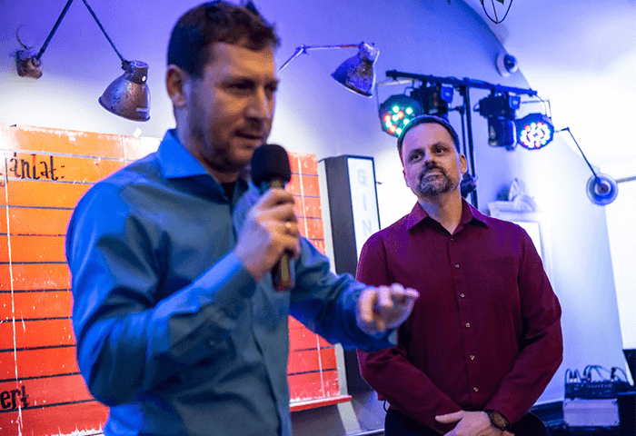 A man in a blue shirt speaking into a microphone, while another man in a red shirt listens attentively in a room with colorful lighting.