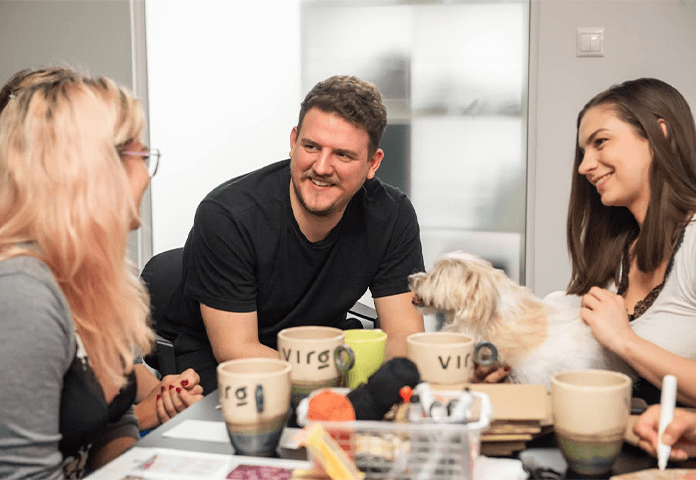 Group of people smiling and chatting around a table with coffee mugs, as a small dog sits on one person's lap.