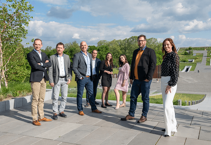 A group of people in business attire standing outdoors on a sunny day, posing on a paved area with greenery and blue skies in the background.