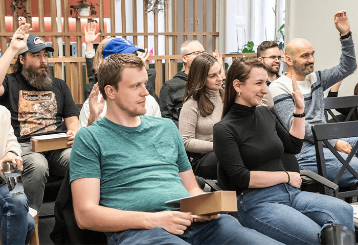 A group of people sitting in a seminar, some with their hands raised, engaged and smiling.