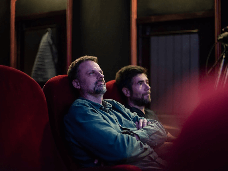 Two men sitting in dark theater seats, watching something attentively.