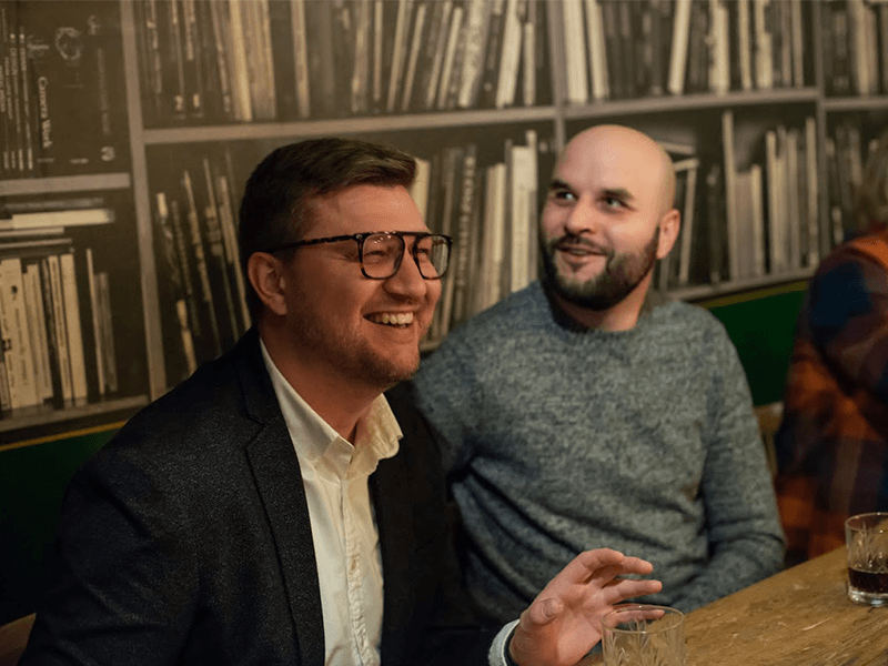 Two men seated at a table, smiling and enjoying a conversation in a cozy setting.