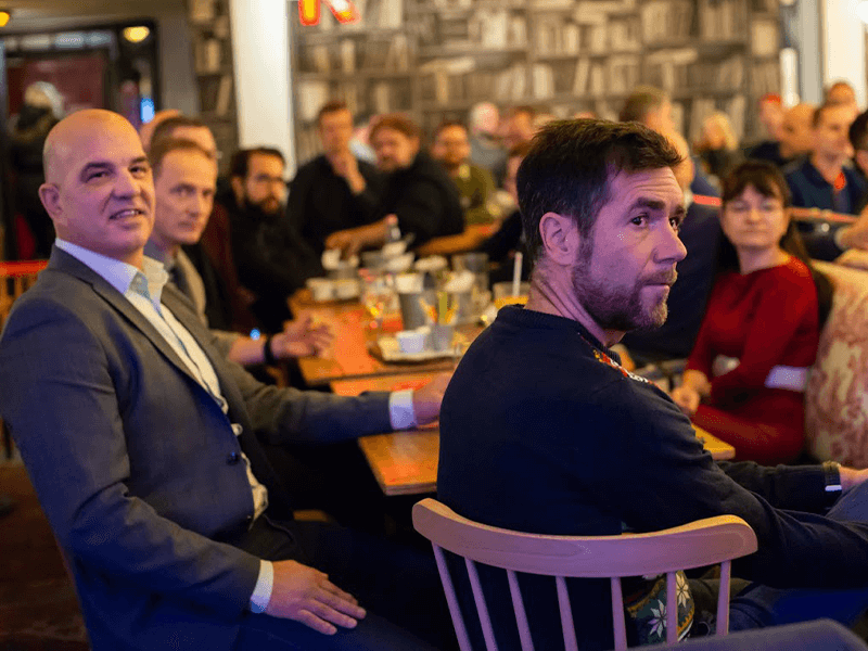 Group of people sitting in a lively, social setting, listening to a presentation.