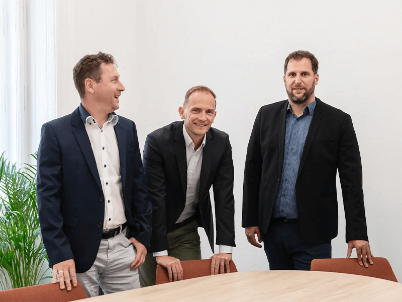 Three men in business attire smiling and standing around a table in a well-lit office