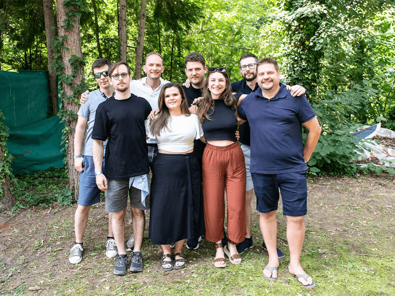 Group of friends smiling and posing together in a green, wooded area.